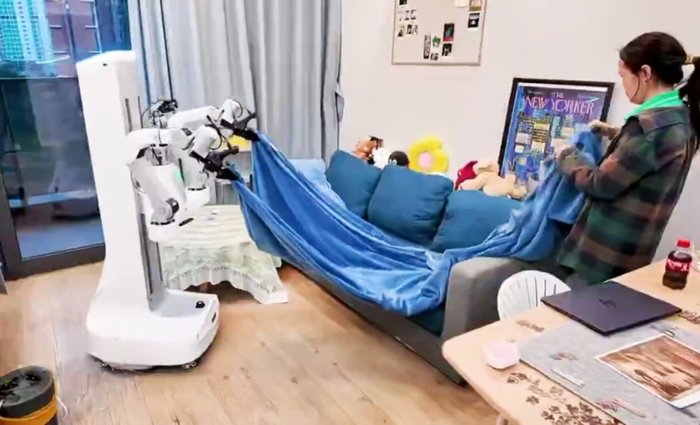 X Square Robot의 Quanta X2 휴머노이드 로봇이 실제 가정에서 소파 위 담요를 정리하는 모습. 옆에서 청소 전문 인력이 함께 협업하고 있다.